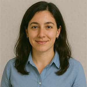 A woman with long hair is smiling while wearing a blue shirt, in front of a wall indoors.