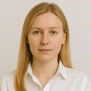 A woman with blonde hair smiling against a wall, captured in a headshot portrait. She is wearing clothing that contrasts with the background, showcasing her neck, chin, and jawline.