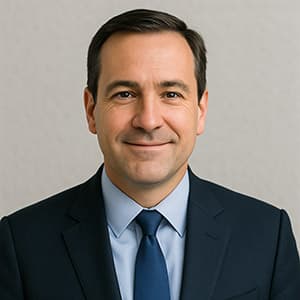 A man in a suit and tie smiling, with a formal appearance and a comb over hairstyle, captured in a headshot. He is indoors against a wall.