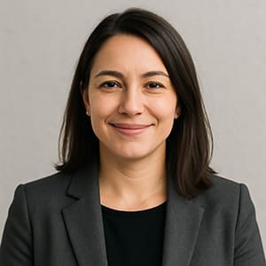 A woman in a suit smiles at the camera, showcasing her face in a headshot. She has well-groomed eyebrows and a confident expression, with indoor fabric textures visible in the background.