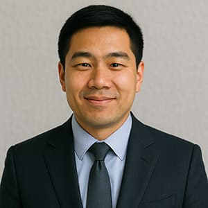 A man wearing a suit and tie smiles for the camera, showcasing a formal appearance with a blazer and dress shirt. His face is visible in the headshot, highlighting features such as his forehead, chin, and eyebrows against an indoor wall background.