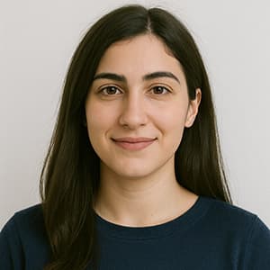 A woman with long hair smiling, standing indoors against a neutral wall, wearing casual clothing.