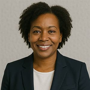 A woman in a suit smiles for the camera, wearing a blazer with a formal design. The background is a simple wall, and her expression conveys confidence and professionalism.